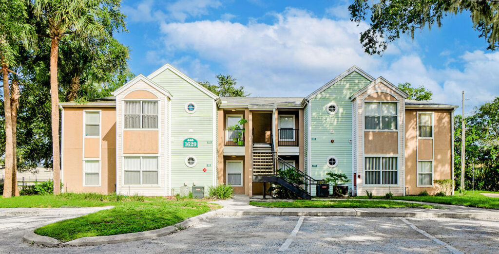 Front view of a two-story multifamily apartment building at Watauga Woods Apartments, featuring light green siding with tan accents, symmetrical gables, and an exterior staircase leading to the second floor, surrounded by palm trees and an empty parking lot.
