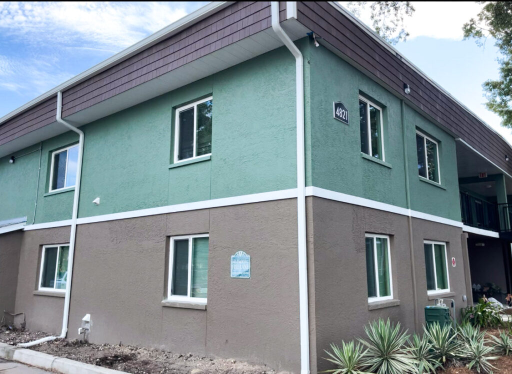 After: Two-story apartment building with a fresh two-tone exterior paint job—green on the upper section and brown on the lower half—accented by white trim and downspouts. The building has several white-framed windows.