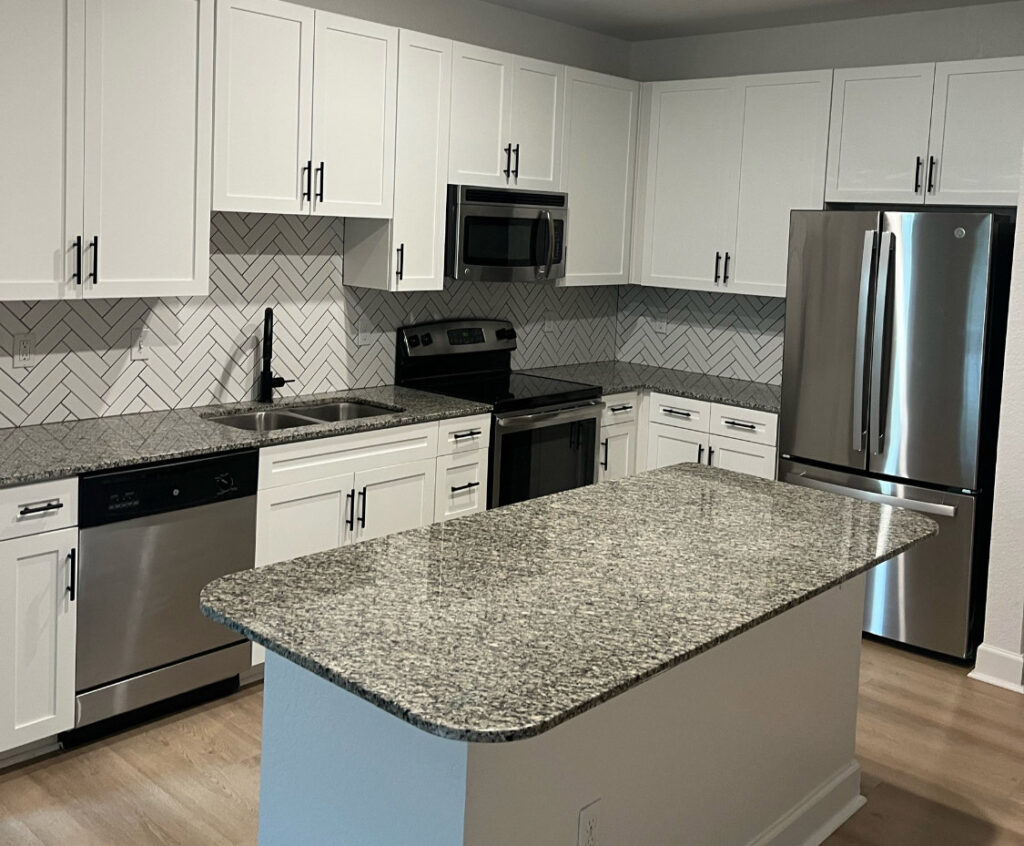 Modern kitchen with white shaker cabinets, granite countertops, and stainless steel appliances, including a French door refrigerator and built-in microwave. A herringbone-patterned backsplash adds texture and contrast behind the black sink and stove, while a central island offers additional prep space.