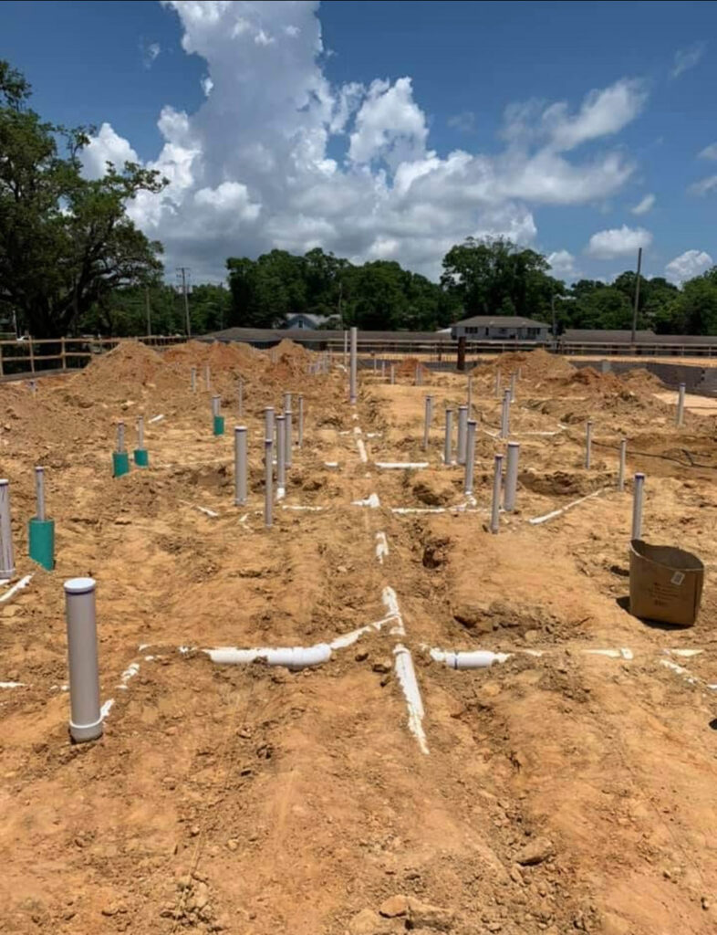 Construction site showing early-stage underground plumbing installation with numerous white PVC pipes and fittings protruding from the dirt foundation. Trenches outline the layout for future utilities, set against a clear sky and surrounded by trees and nearby buildings.