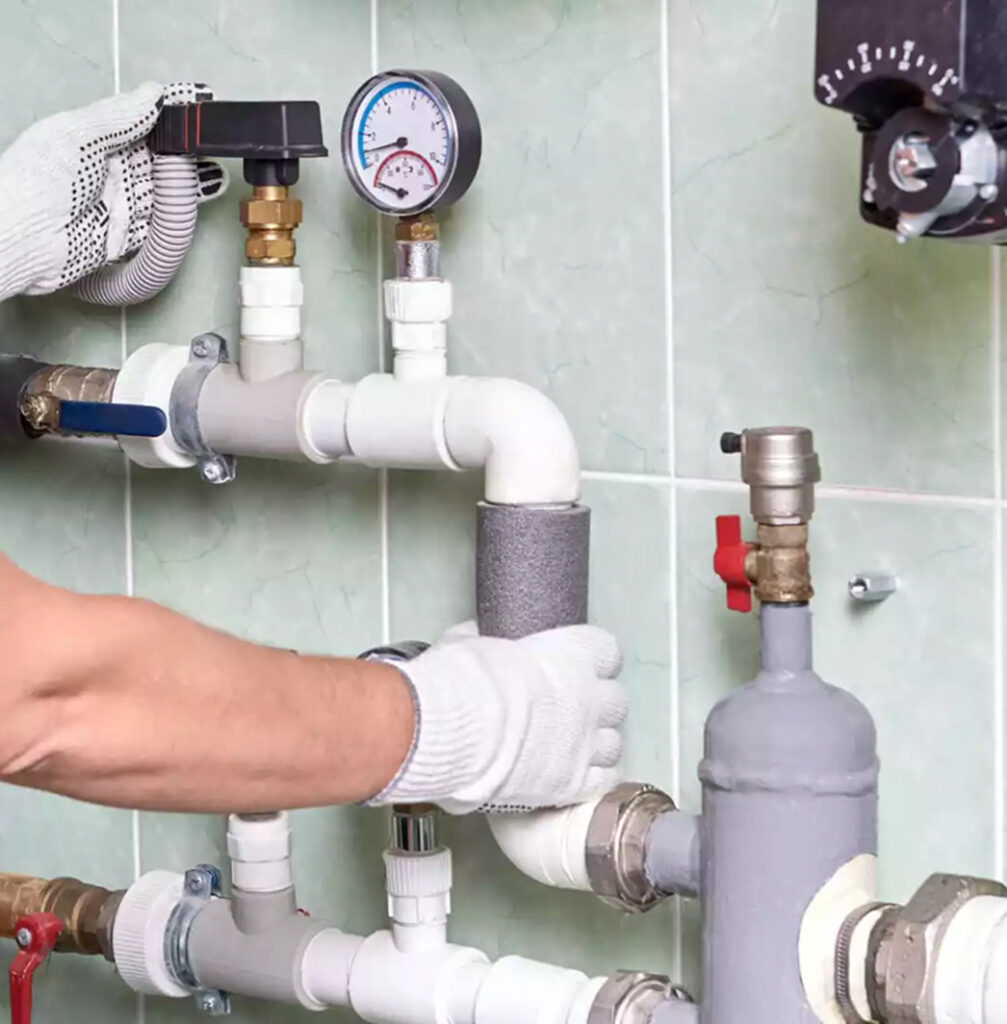 Close-up of a plumber’s gloved hands adjusting or installing insulated piping in a mechanical room. The setup includes various white PVC and metal pipes, valves, and a pressure gauge mounted on a tiled wall.