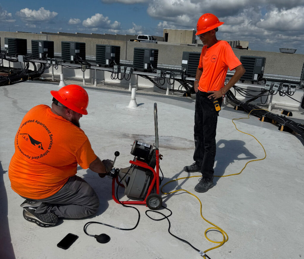 Two HVAC technicians wearing orange shirts and red hard hats work on a rooftop surrounded by air conditioning units. One kneels beside a portable vacuum pump or testing machine, while the other stands observing with a tool clipped to his pocket, under a sunny sky with scattered clouds.