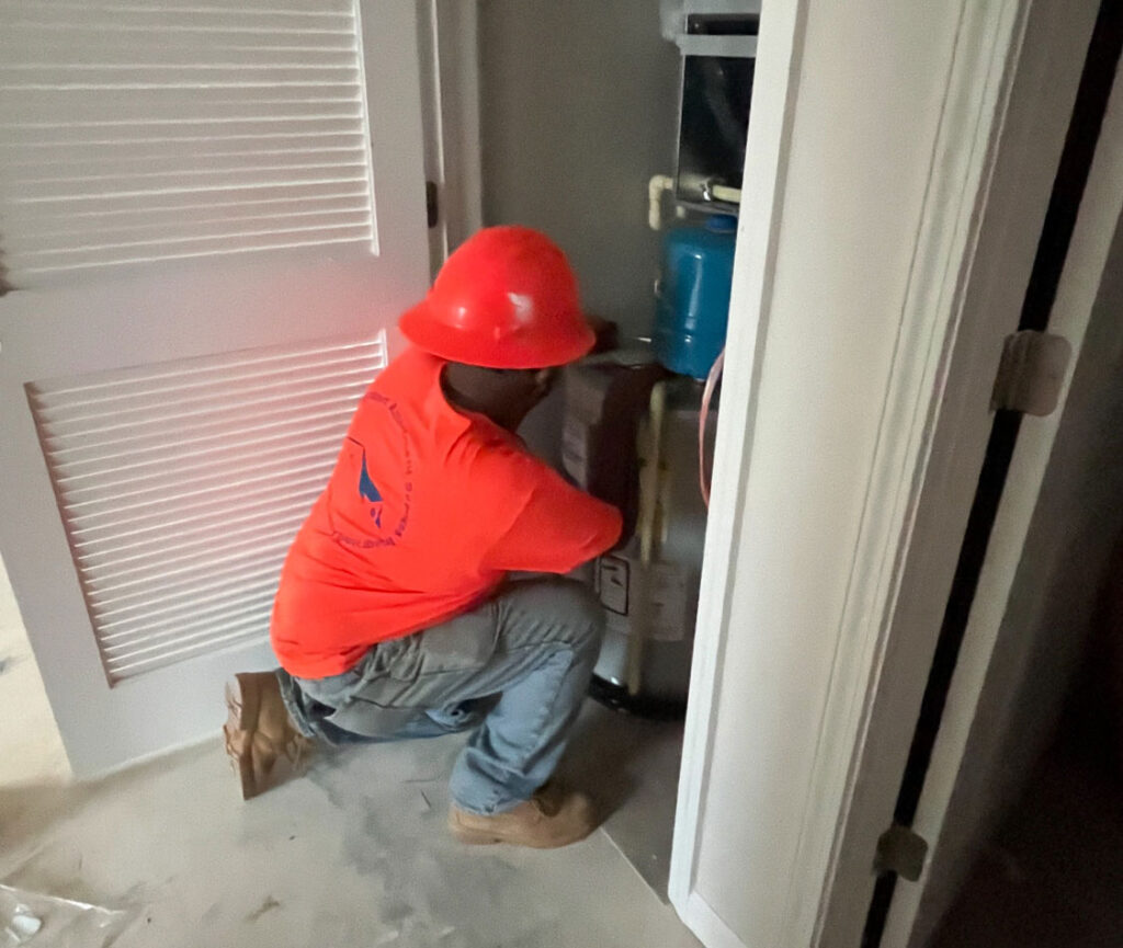 A construction worker wearing a red hard hat and neon orange shirt is crouched in front of a water heater or HVAC system inside a closet, appearing to inspect or service the unit. The setting suggests an indoor maintenance or installation task in a residential or commercial building.