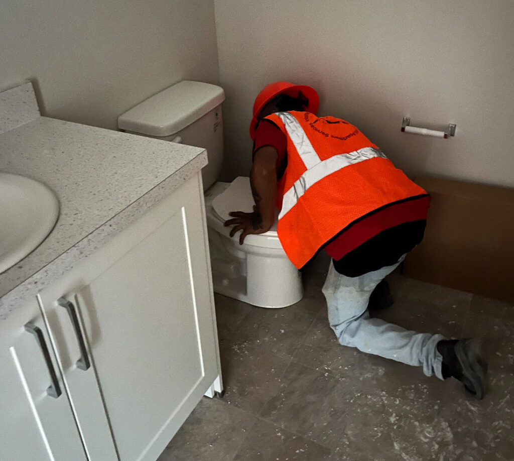 A construction worker wearing a safety vest and hard hat kneels on the bathroom floor while inspecting or installing a toilet next to a white vanity. The space appears to be under construction or newly renovated, with no toilet paper on the holder and minimal fixtures.